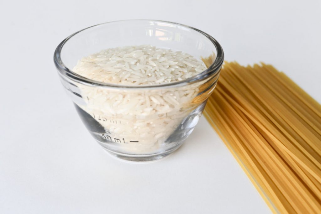 Food portion control. Measuring bowl filled with uncooked rice and dry spaghetti on a table next to it