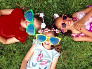 Three girls laying on the grass with giant sunglasses on and smiling