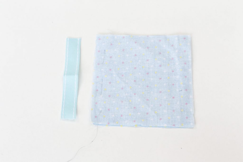 A piece of blue 1cm wide ribbon 10cm long is laid on the table perpendicular to the squares of blue spotted fabric that have been stitched along one seam