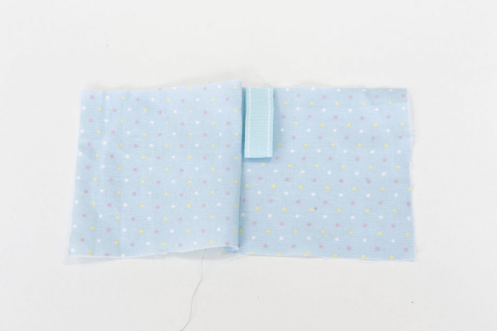 After stitching together the two pieces of blue spotted fabric along the first side. The fabric is opened out flat and a piece of 10cm ribbon, folded in half to form a loop is places along the seem with the open edge lined up with the second side of the fabric