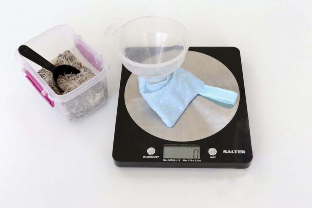 The pyramid lavender bag in blue material with spots on a set of scales with a plastic funnel inside so that the rice/lavender mixt (pictured in a container) can be spooned in until the pre-zeroes weight reaches approximately 80g