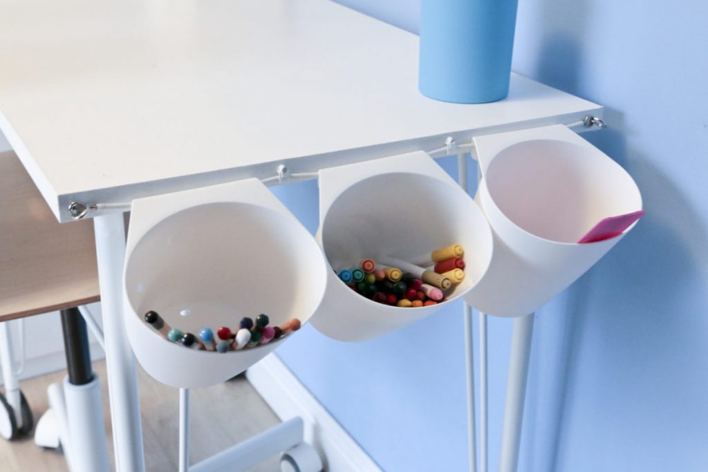 Close-up of three Ikea Bygel containers hanging on a net curtain wire on the side of a children's white desk filled with pens and pencils