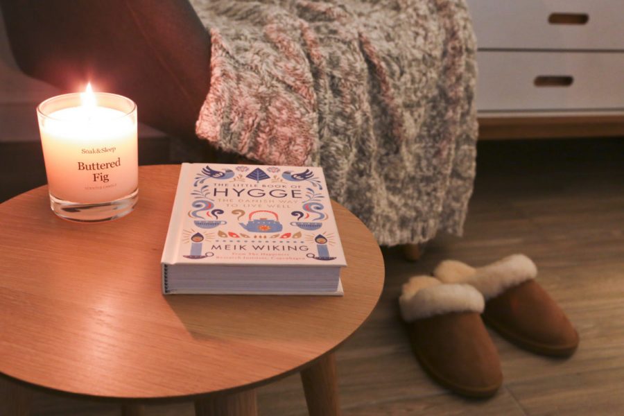 Candle and Hygge book on side table near an armchair with cable knit throw and slippers on floor