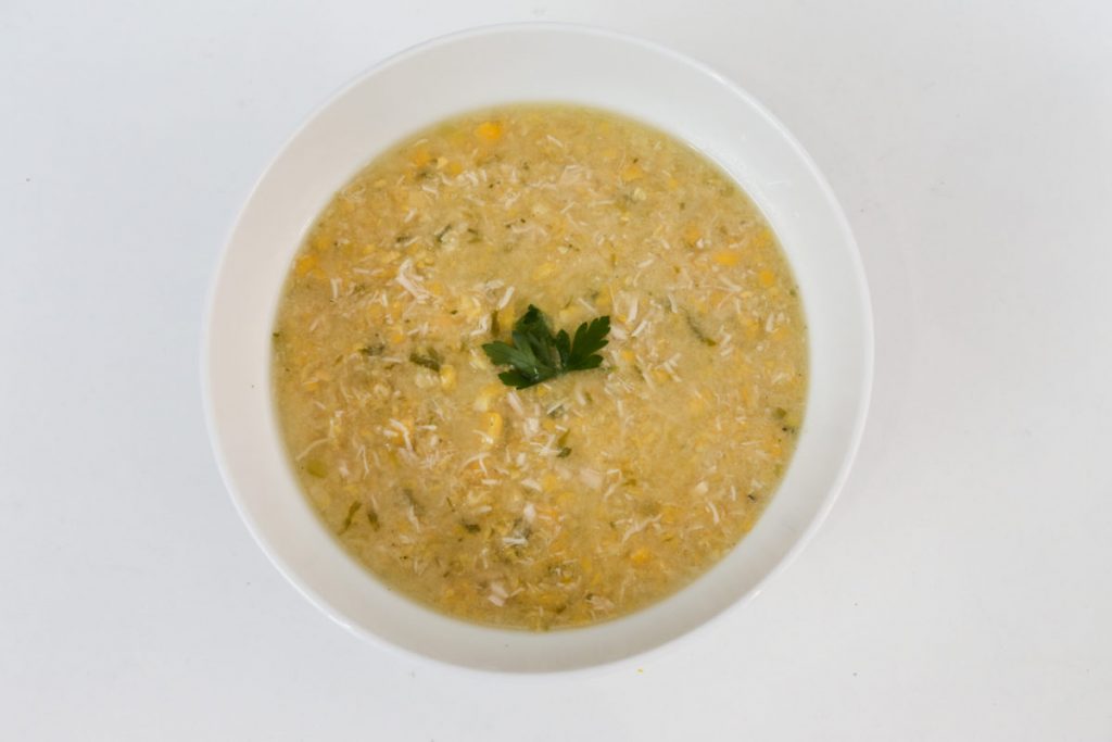 Chunky Chicken & Sweetcorn Soup in a white bowl on a white table with a sprig of parsley on top.