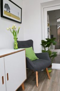 Wayfair Pantone Greenery inspiration close-up of grey chair with green cushion next to a table with a green vase containing tulips and an artificial floor standing Taro plant